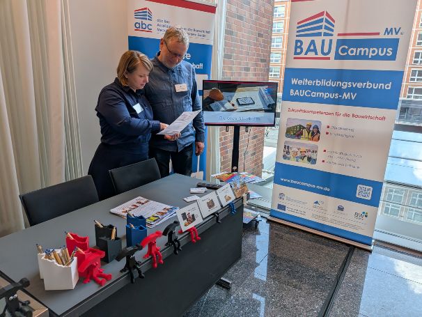 Eine Frau steht an ihrem Messestand und erklärt einem Interessierten etwas