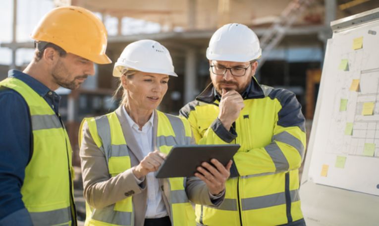 Drei Fachkräfte der Bauwirtschaft besprechen Veränderungen und neue Prozesse auf einer Baustelle. Eine Führungskraft erläutert die nächsten Schritte anhand eines Tablets.