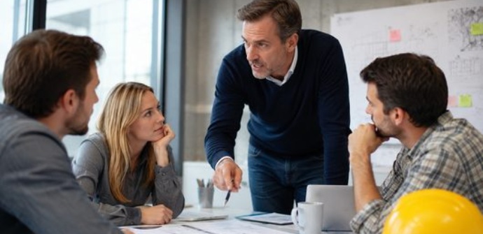 Realistische Büroszene aus einem Baubüro: Ein Team bespricht Unterlagen und Baupläne, während kommunikative Dynamiken und Machtmittel im Arbeitsalltag sichtbar werden.