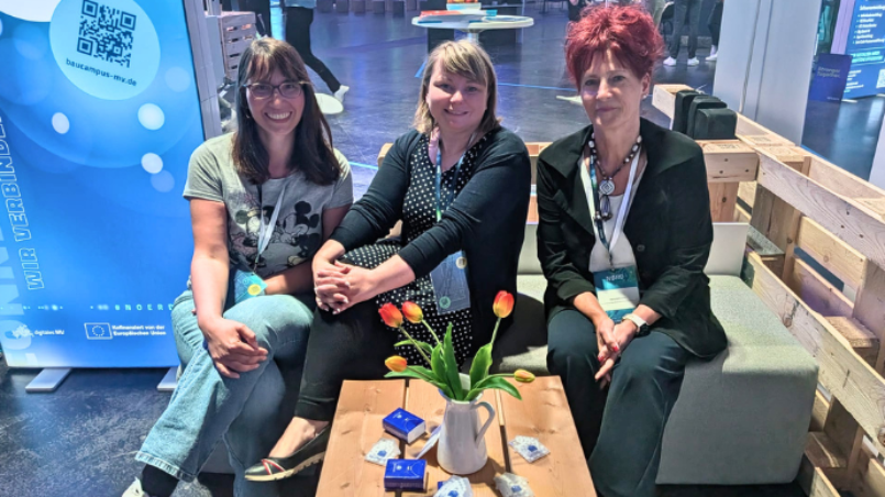 Drei Vertreterinnen des BAUCampus MV sitzen an einem Messestand auf der NØRD 2026. Vor ihnen steht ein Tisch mit Blumen, im Hintergrund sind das BAUCampus-MV-Rollup und die Messeumgebung zu sehen.